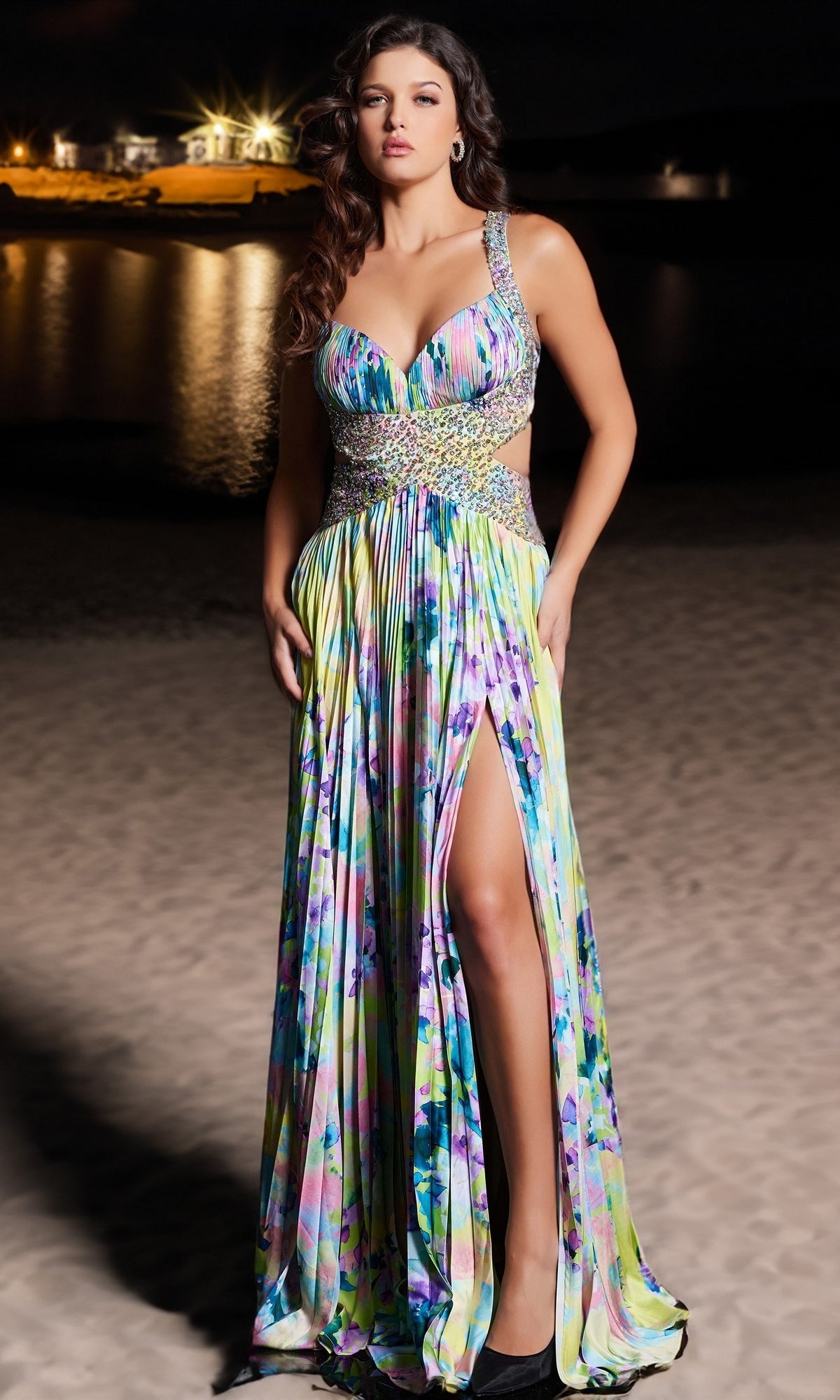 A woman stands on a sandy beach at night wearing the Long Prom Dress 38733 by Jovani, a colorful floral beaded dress with a thigh-high slit and jeweled details. Lights from distant buildings reflect on the water behind her.